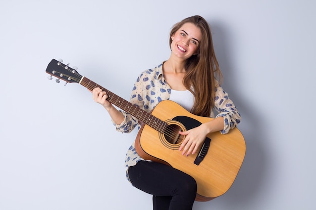 Gelukkige jonge vrouw in beige blouse en zwarte broek die een gitaar op grijze achtergrond in studio houdt