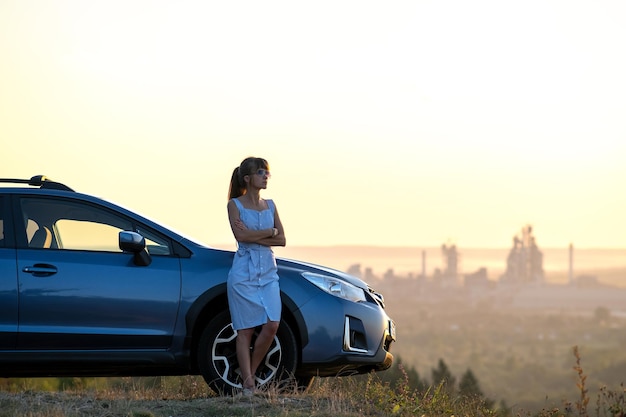 Gelukkige jonge vrouw in blauwe jurk die in de buurt van haar voertuig staat en kijkt naar de zonsondergang van de zomerse natuur. Reizen en vakantie concept.