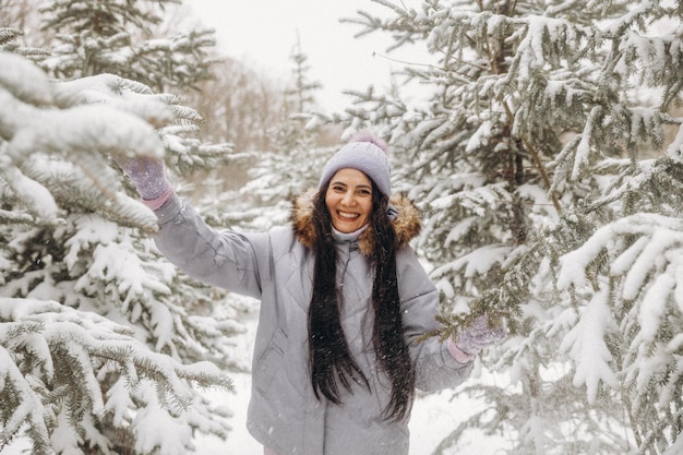 Gelukkige jonge vrouw in de winter op een wandeling in de natuur. een vrouw in een lila jas staat bij de kerstbomen in een winterpark.