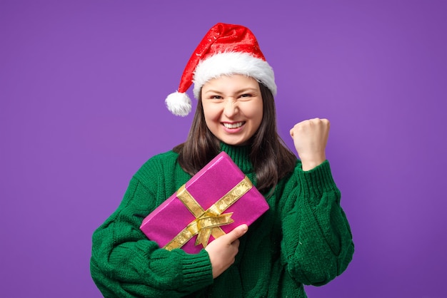 Gelukkige jonge vrouw in Kerstmisglb met gift op geïsoleerd paars