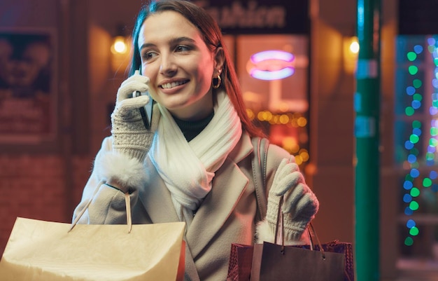 Foto gelukkige jonge vrouw loopt door de straten van de stad en doet kerstinkopen ze draagt veel tassen en heeft een telefoontje met haar smartphone