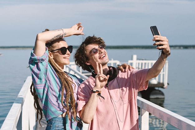 Gelukkige jonge vrouw met dreadlocks en haar vriendje in zalmhemd dat op de pier staat en selfie neemt voor sociale media