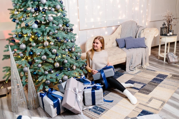 Gelukkige jonge vrouw met een cadeau in de buurt van de kerstboom witte zilveren kleur op de achtergrond van de bank prettige kerstdagen en nieuwjaar sfeervolle momenten