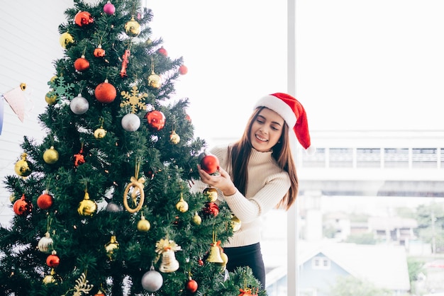 Gelukkige jonge vrouw met een kerstmuts en trui glimlach en geniet van het versieren van verschillende kerstballen op de kerstboom in de woonkamer thuis Genieten en vieren van de kerstvakantie