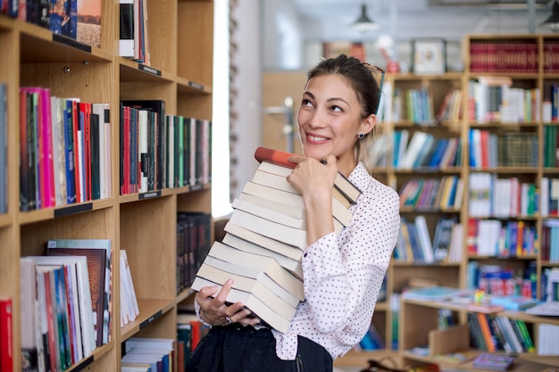 Gelukkige jonge vrouw met een stapel boeken in bibliotheek