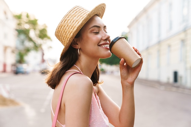 gelukkige jonge vrouw met een zomerse strohoed die lacht en koffie drinkt terwijl ze op straat in de stad loopt