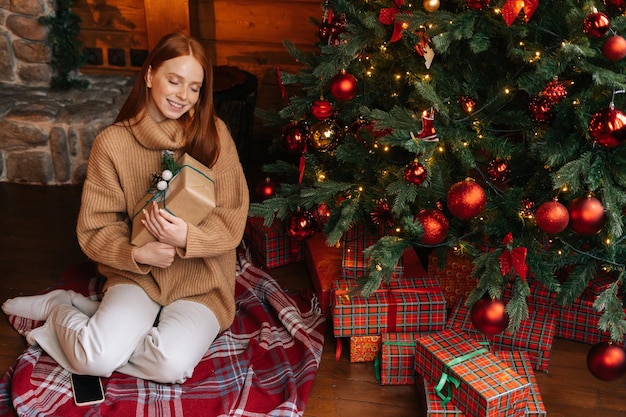 Gelukkige jonge vrouw met geschenkdoos ontvangen voor Kerstmis zittend op de vloer achtergrond van kerstboom