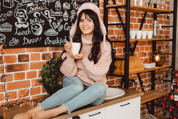 Gelukkige jonge vrouw met lang haar en in een winterbontmuts drinkt koffie in de keuken thuis. kerst concept.