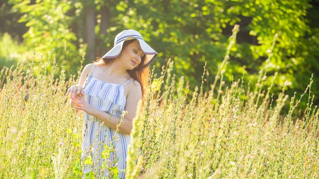 Gelukkige jonge vrouw met lang haar in hoed en jurk trekt haar handen naar de planten terwijl ze op een zonnige dag door het zomerbos loopt. Banner met kopieerruimte