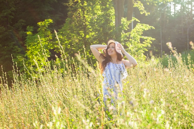 Gelukkige jonge vrouw met lang haar in hoed en jurk trekt haar handen naar de planten terwijl ze op een zonnige dag door het zomerbos loopt. Zomer vreugde