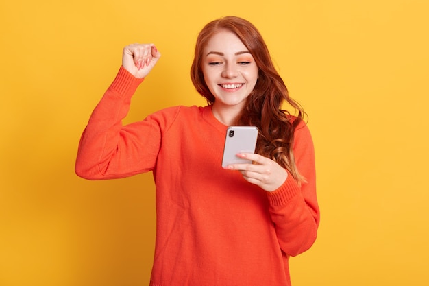 Gelukkige jonge vrouw met mooi lang rood golvend haar, schreeuwend en balde vuist terwijl ze mobiele telefoon in de hand houdt, poseren geïsoleerd over gele muur