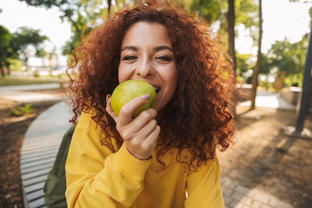 Gelukkige jonge vrouw met rood krullend haar zittend op een bankje in het park, groene appel bijtend
