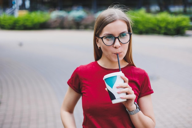 Gelukkige jonge vrouw met rugzak glimlachend en wegkijkend terwijl ze op straat in de stad loopt en geniet van verse koffie om mee te nemen