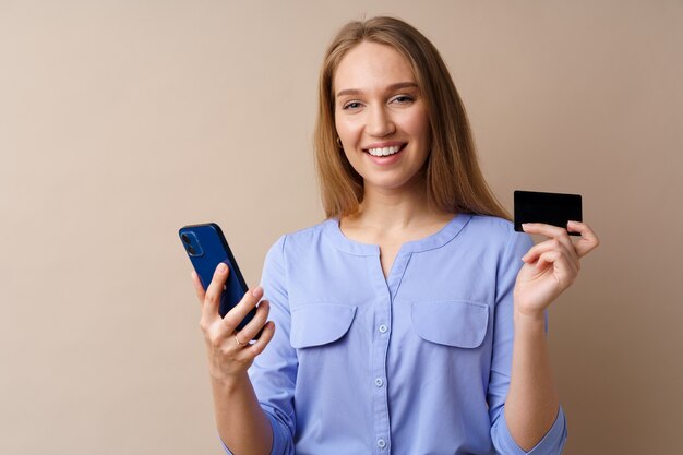 Gelukkige jonge vrouw met smartphone en creditcard tegen beige achtergrond