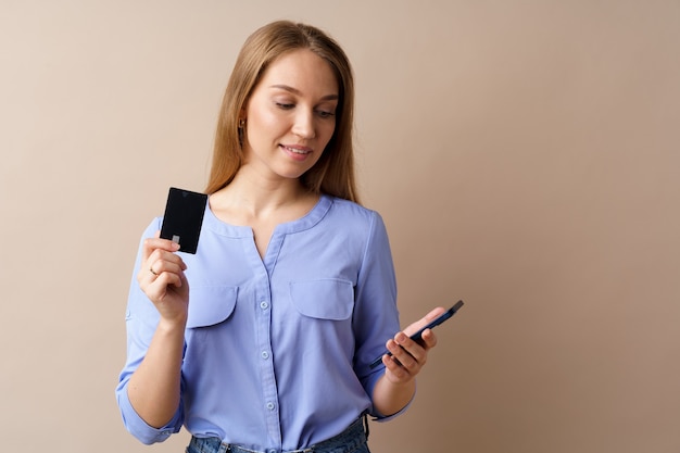 Gelukkige jonge vrouw met smartphone en creditcard tegen beige achtergrond