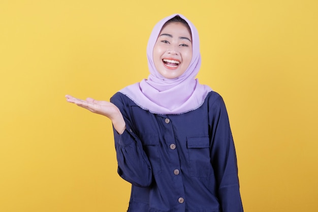 Gelukkige jonge vrouw met vrolijke gezichtsuitdrukking toont iets in haar hand met hijab en vrijetijdskleding in geel