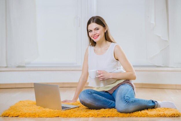 Gelukkige jonge vrouw of student met laptop die thuis op de vloer zit