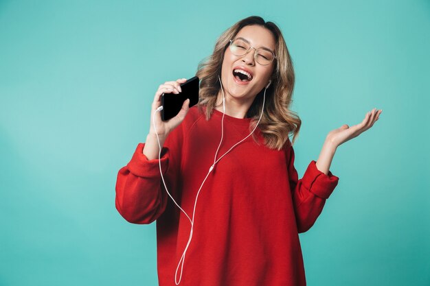 Gelukkige jonge vrouw poseren geïsoleerd over blauwe muur muur luisteren naar muziek met koptelefoon met behulp van mobiele telefoon