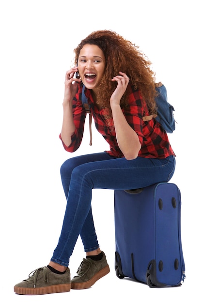 Gelukkige jonge vrouw praat op de mobiele telefoon terwijl ze op een koffer zit op een geïsoleerde witte achtergrond