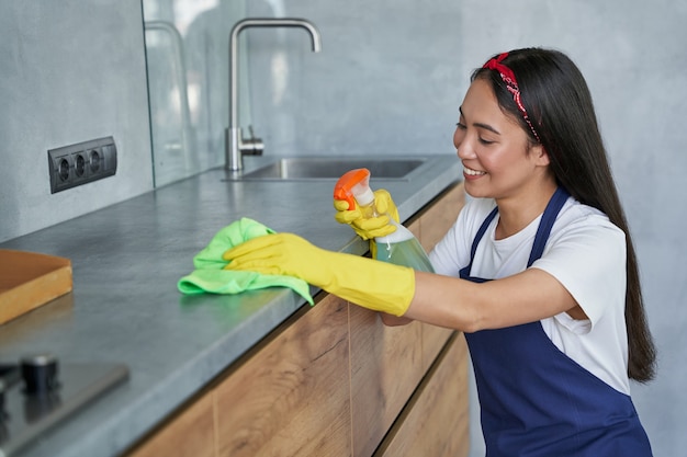 Gelukkige jonge vrouw, schoonmaakster die lacht terwijl ze de keuken schoonmaakt en de oppervlakken besproeit met wasmiddel uit een spuitfles. huishoudelijk werk en huishouden, schoonmaakserviceconcept