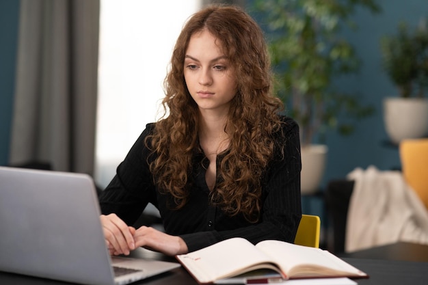 Gelukkige jonge vrouw zit aan het bureau en kijkt naar het scherm van de laptop terwijl ze thuis surft op draadloos internet