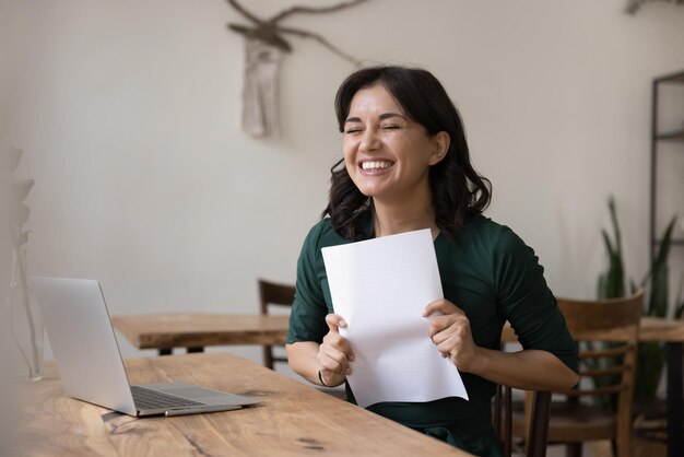 Gelukkige jonge vrouw zitten thuis werkplaats houden papier brief ontvangen bank lening goedkeuring kennisgeving baan promotie of salarisverhoging verrast door groot nieuws geld terugbetaling document genieten moment van succes