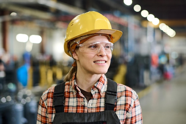 Gelukkige jonge vrouwelijke ingenieur in veiligheidshelm en overall die in de werkplaats staat