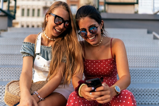 Gelukkige jonge vrouwelijke vrienden in zonnebrillen zitten en surfen op internet op de mobiele telefoon van de stadsstraat