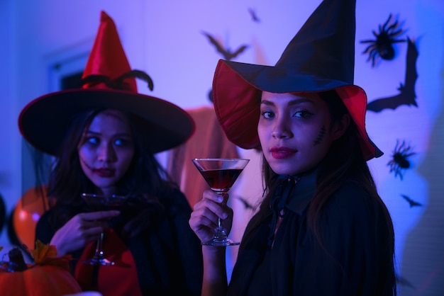 Gelukkige jonge vrouwen in kostuums van zwarte hekshalloween in een partij met pompoen en cocktails