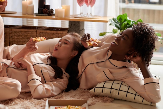 Gelukkige jonge vrouwen in pyjama's die op de vloer rusten, op elkaar leunen en plakjes delicio eten