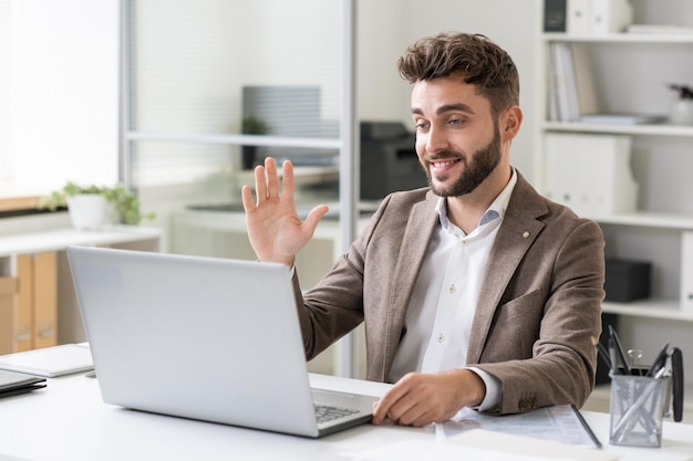 Gelukkige jonge zakenman die hand zwaait naar iemand op het scherm van de laptop