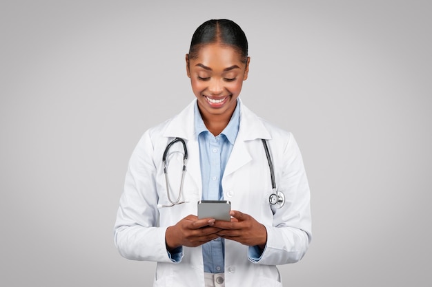 Gelukkige jonge zwarte vrouwelijke dokter in witte jas die aan het typen is bij mobiele telefoonwerk