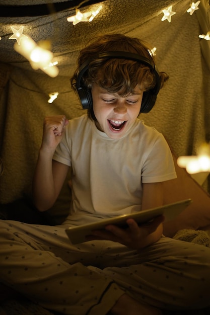 Gelukkige jongen in een headset met een headset die interessante video's kijkt op een moderne tablet terwijl hij in een gezellige kamer naast een gloeiende krans zit