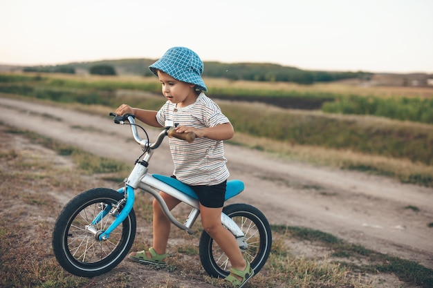 Gelukkige jongen met een fiets en hoed loopt in een veld in een zomeravond en probeert een kleine heuvel te rijden