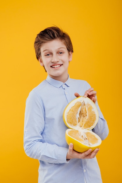 Gelukkige jongen met pompelmoesfruit in hand, exotisch fruit, banaan en sinaasappel, in het blauwe overhemd, dat op gele muur wordt geïsoleerd