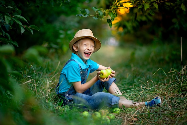 Gelukkige jongen van zes jaar zit op het gras in een appelboomgaard en houdt een appel in zijn handen.