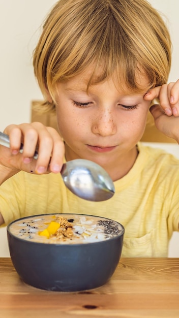 Gelukkige jongen zit en eet een verse smoothiekom met mango-drakenfruitgranola en chiazaden