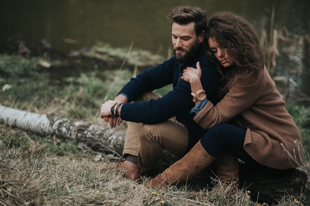 Gelukkige Kaukasische minnaars zitten aan de oever van het meer. Jong koppel is knuffelen op herfstdag buitenshuis. Een bebaarde man en een gekrulde vrouw verliefd. Concept van liefde en familie.