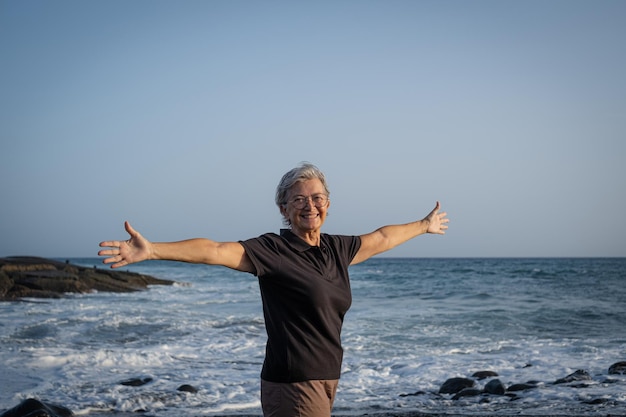 Gelukkige Kaukasische senior vrouw staande op het strand bij zonsondergang met uitgestrekte armen oudere vrouw genieten van vrije tijd vakantie of pensioen