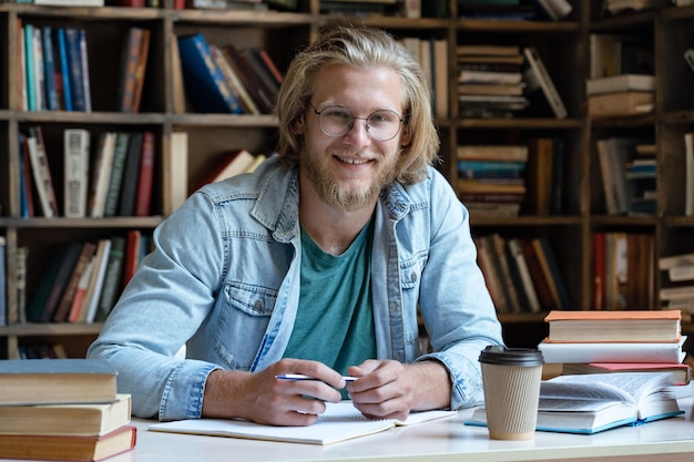 Gelukkige kerel studentenstudie in bibliotheek kijkend naar camera zit aan bureau