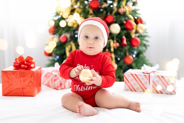 Gelukkige kindbaby in rood kerstkostuum die nieuwjaar thuis viert met kerstboom en geschenken
