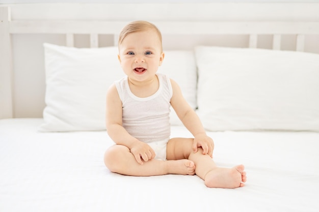 Foto gelukkige kleine baby meisje of jongen in een wit bodice zit op het bed in de slaapkamer en glimlacht of lacht levensstijl