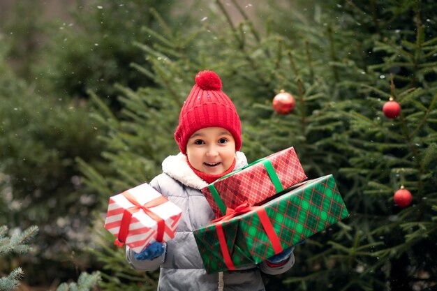 Gelukkige kleine glimlachende meisje in rode pet met kerstcadeau dozen staan in de buurt van sparren