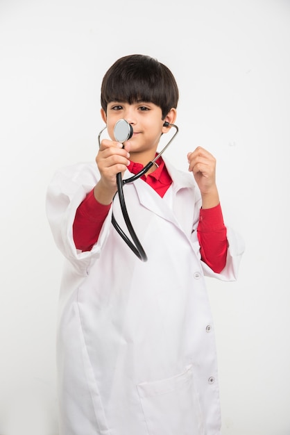 Gelukkige kleine Indiase Aziatische jongen in medisch uniform als arts, stethoscoop vasthoudend en kijkend naar camera geïsoleerd op witte achtergrond