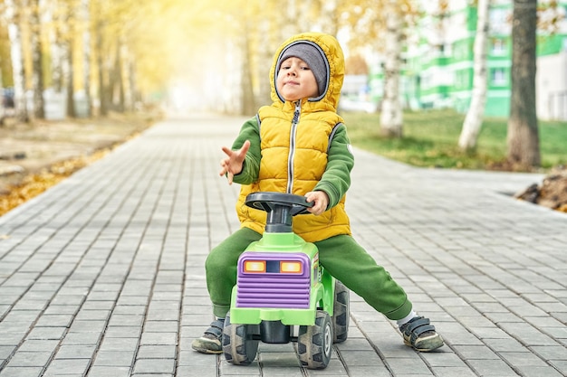 Gelukkige kleine jongen kind in heldergeel vest duwt rideon speelgoed terwijl het lopen op geplaveide weg in de herfst