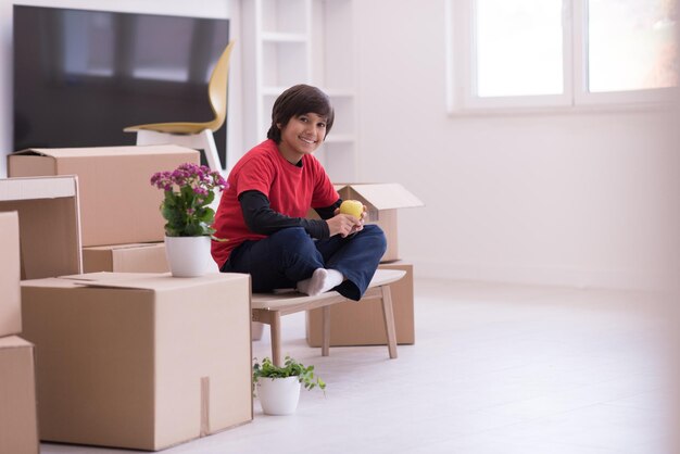 gelukkige kleine jongen zittend op de tafel met kartonnen dozen om hem heen in een nieuw modern huis