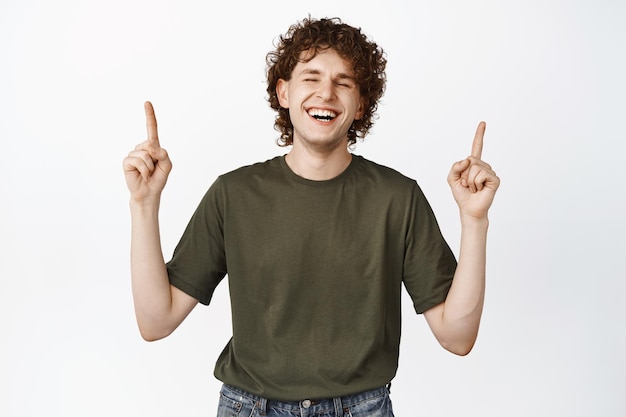 Gelukkige knappe jongeman met krullend haar die met de vingers omhoog wijst en lacht en reclame toont met een witte achtergrond voor een promo-deal