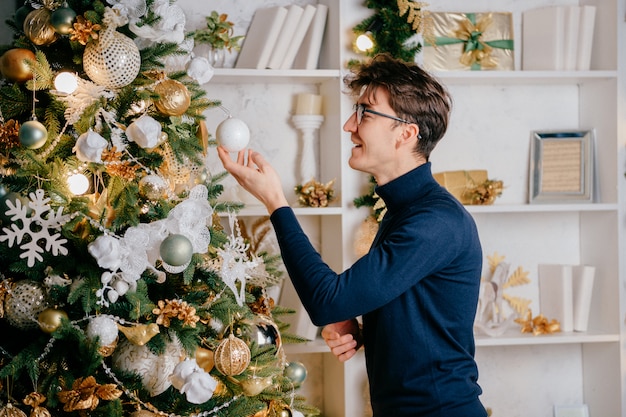 Gelukkige knappe mens die cristmasboom in comfortabele ruimte met de nieuwe decoratie van de jaarvakantie verfraaien.