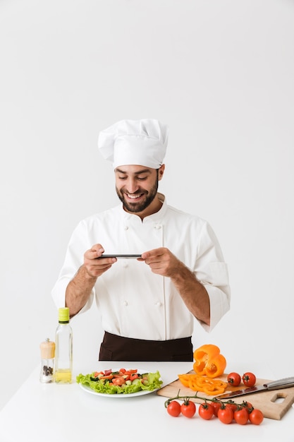 gelukkige kok man in uniform glimlachend en het nemen van foto van bord met groentesalade op smartphone geïsoleerd over witte muur