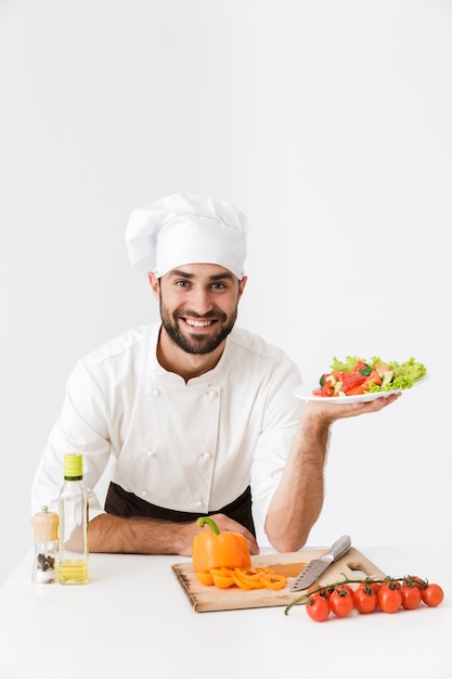 gelukkige kok man in uniform glimlachend en vasthoudend bord met groentesalade geïsoleerd over witte muur